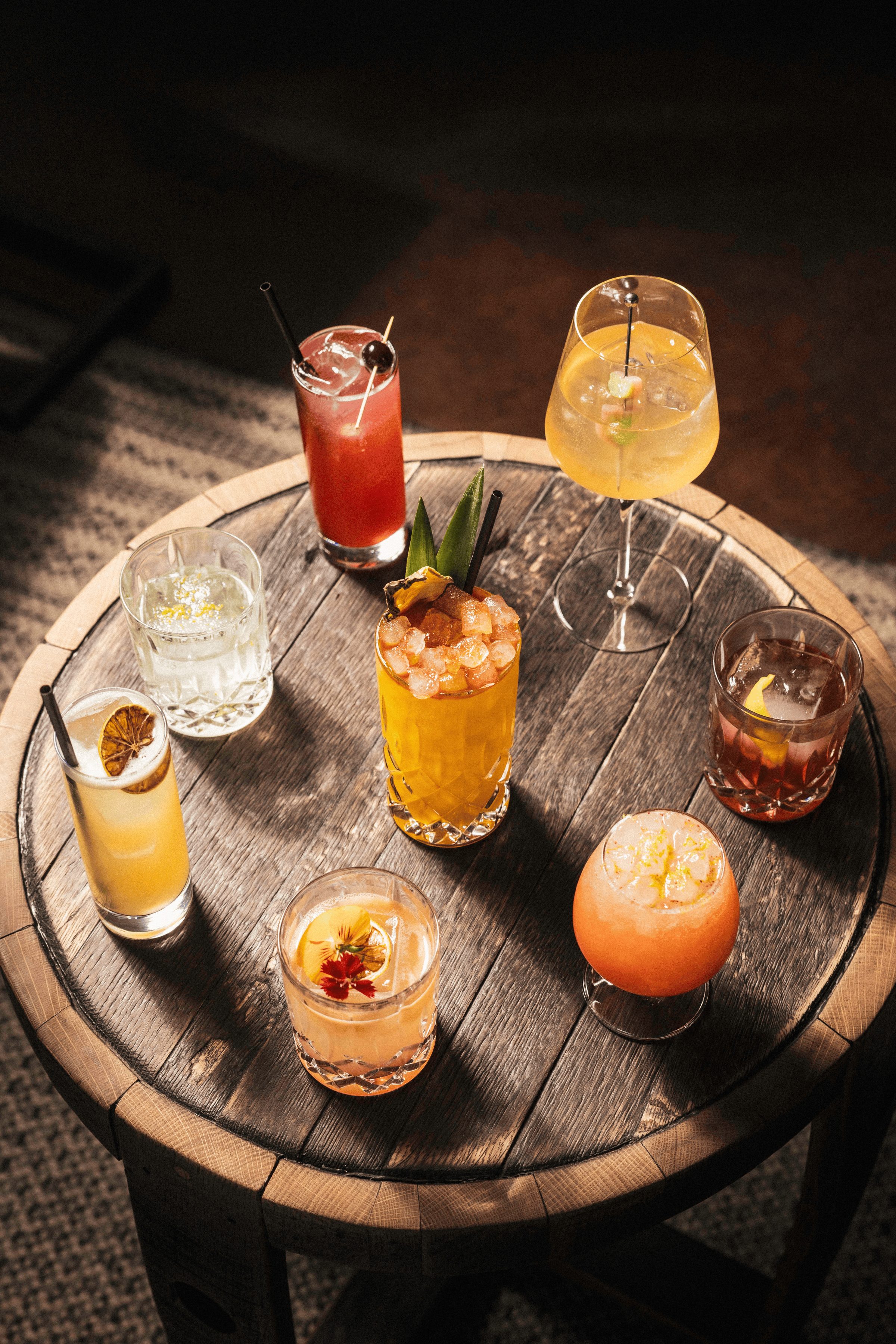 Eight vibrant cocktails, each unique in color and garnish, are beautifully arranged on a rustic wooden table.