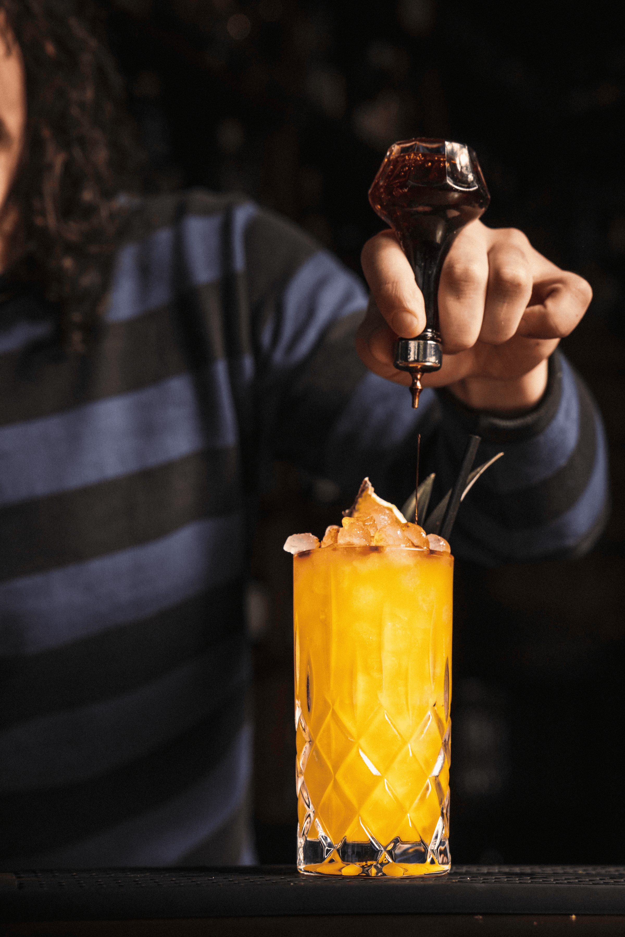 A bartender skillfully drizzles dark syrup over a vibrant orange cocktail, garnished with crushed ice and stylish green leaves.